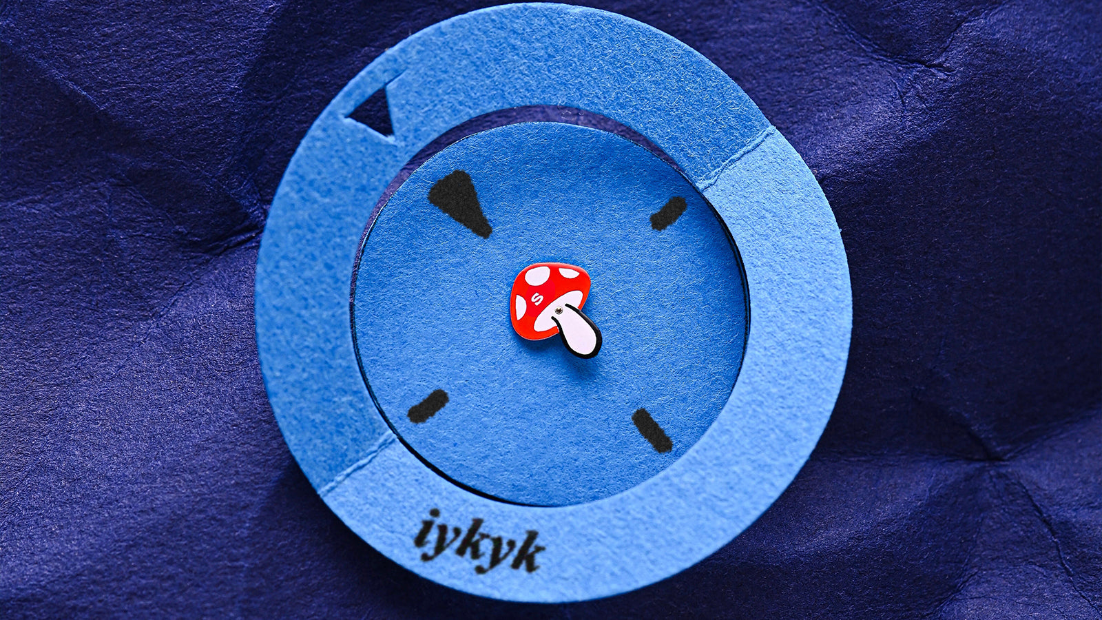 A macro photo of the paper art representation of the Q Timex watch face with blue circular frame, red mushroom seconds hand, and “iykyk” text on blue crumpled paper.
