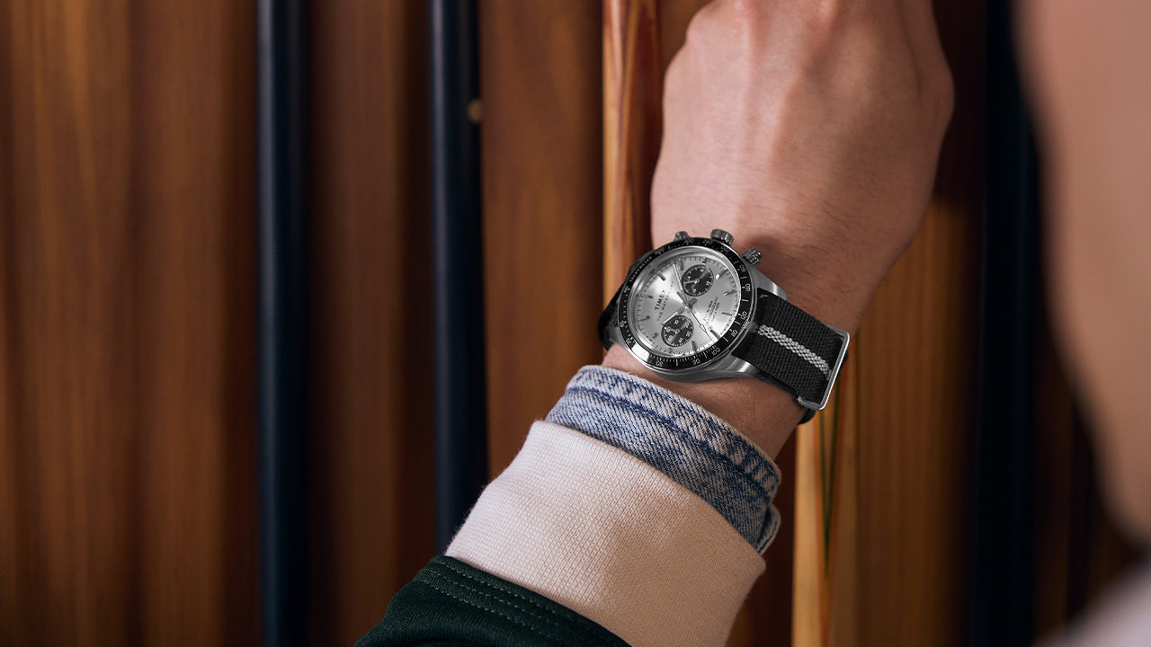 Person wearing the Timex Waterbury Heritage Chronograph with silver and black dial and striped fabric strap while holding a pool cue near a wooden rack.