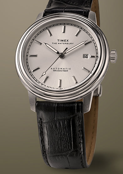 Timex Waterbury Metropolitan Automatic watch with a silver sunray dial, polished stainless steel case, and black leather strap on a beige background.