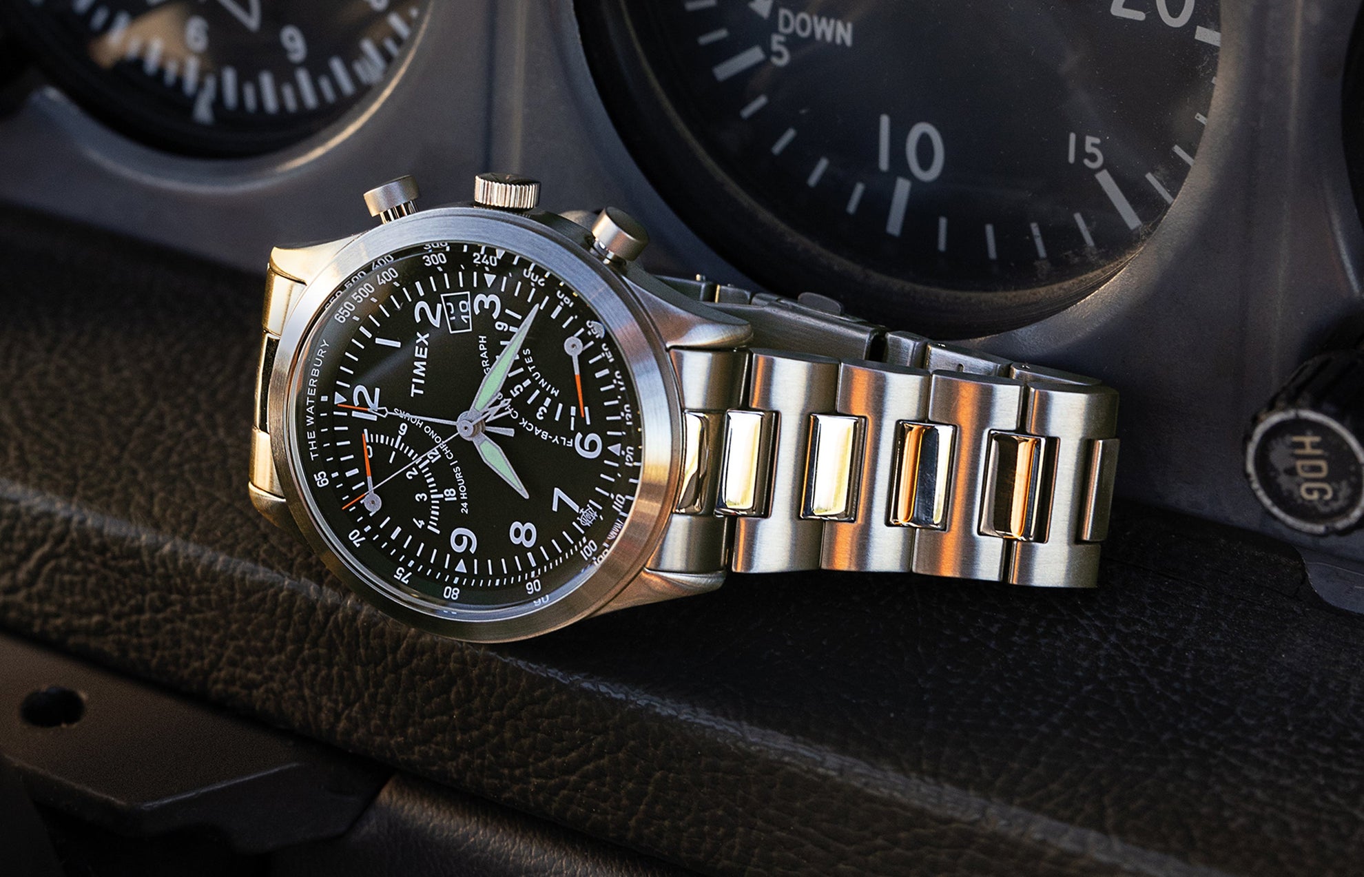 Timex Waterbury Traditional Fly-Back Chronograph resting on an aircraft instrument panel, featuring a black dial, stainless steel bracelet, and luminous hands.