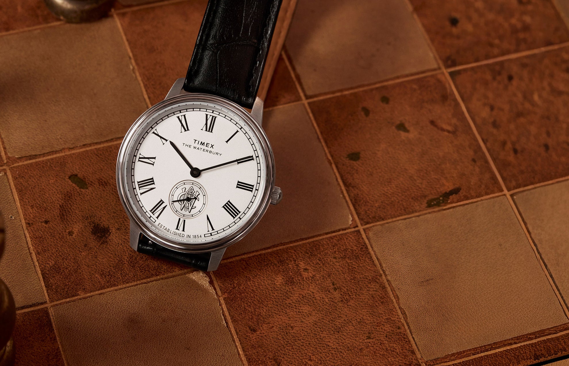 Timex Waterbury Metropolitan Sub-Second watch with white dial, Roman numerals, and black leather strap resting on a vintage chessboard.
