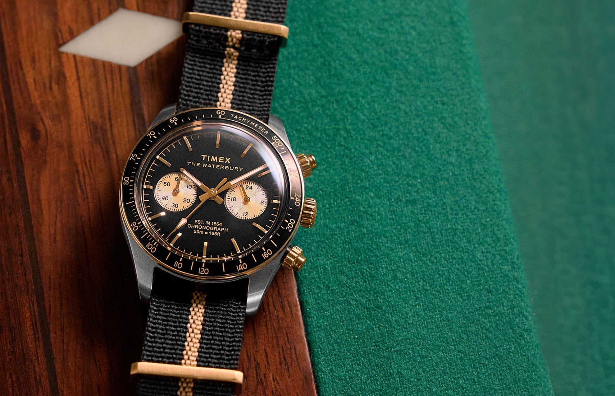 Timex Waterbury Heritage Chronograph with black dial, gold-tone accents, and black fabric strap featuring a tan center stripe resting on a wooden pool table.