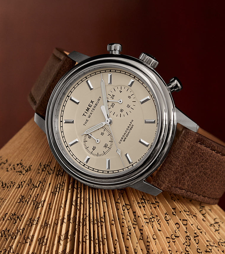 Timex Waterbury Metropolitan Chronograph with cream dial, stainless steel case, and brown leather strap resting on an open book with folded pages.