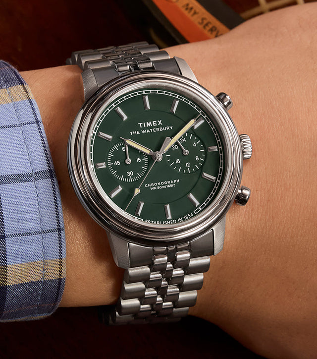 Timex Waterbury Metropolitan Chronograph with green dial and stainless steel bracelet worn on wrist, paired with a plaid shirt and classic styling.