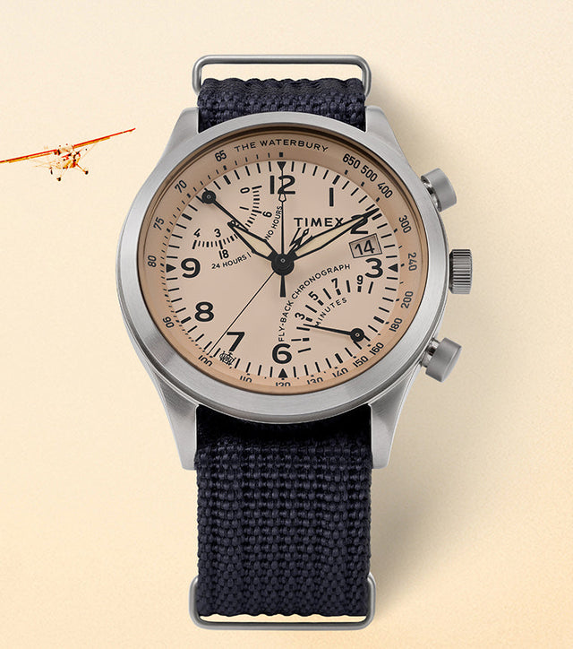 Timex Waterbury Traditional Fly-Back Chronograph with stainless-steel case, beige aviation-style dial, and navy fabric strap displayed on a cream background with a vintage plane in view.