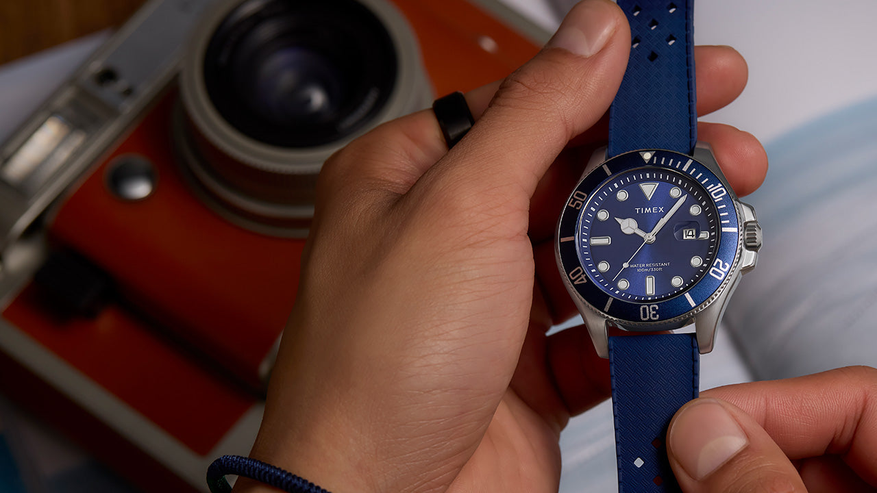 Close-up of a person holding a Timex Harborside Coast watch with a blue dial and bezel, and blue textured strap, near an orange vintage camera.