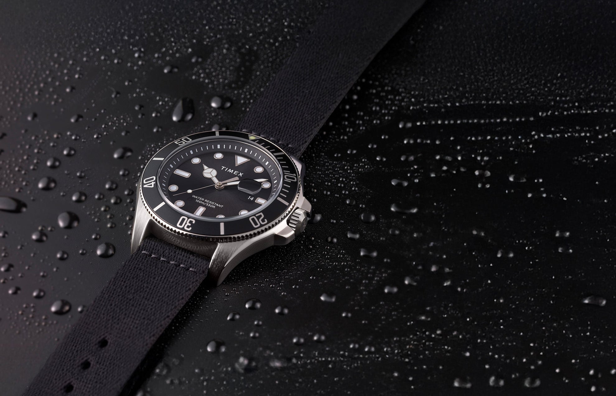 TIMEX® Harborside Coast watch with black dial and black fabric strap resting on a water-dappled black surface, showcasing its water-resistant design.
