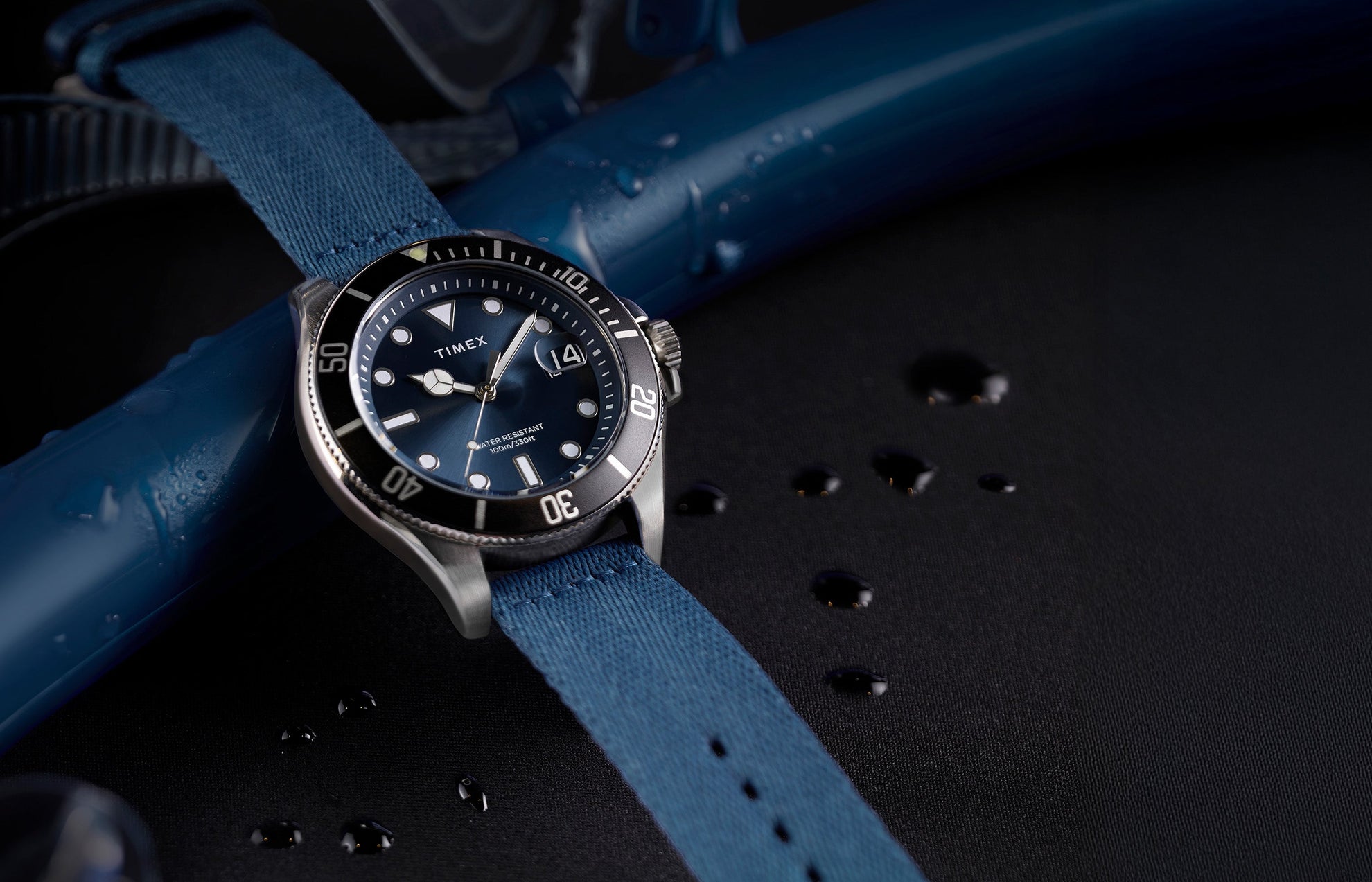 TIMEX® Harborside Coast watch with blue dial, silver-tone case, and blue fabric strap resting on a wet surface with water droplets.