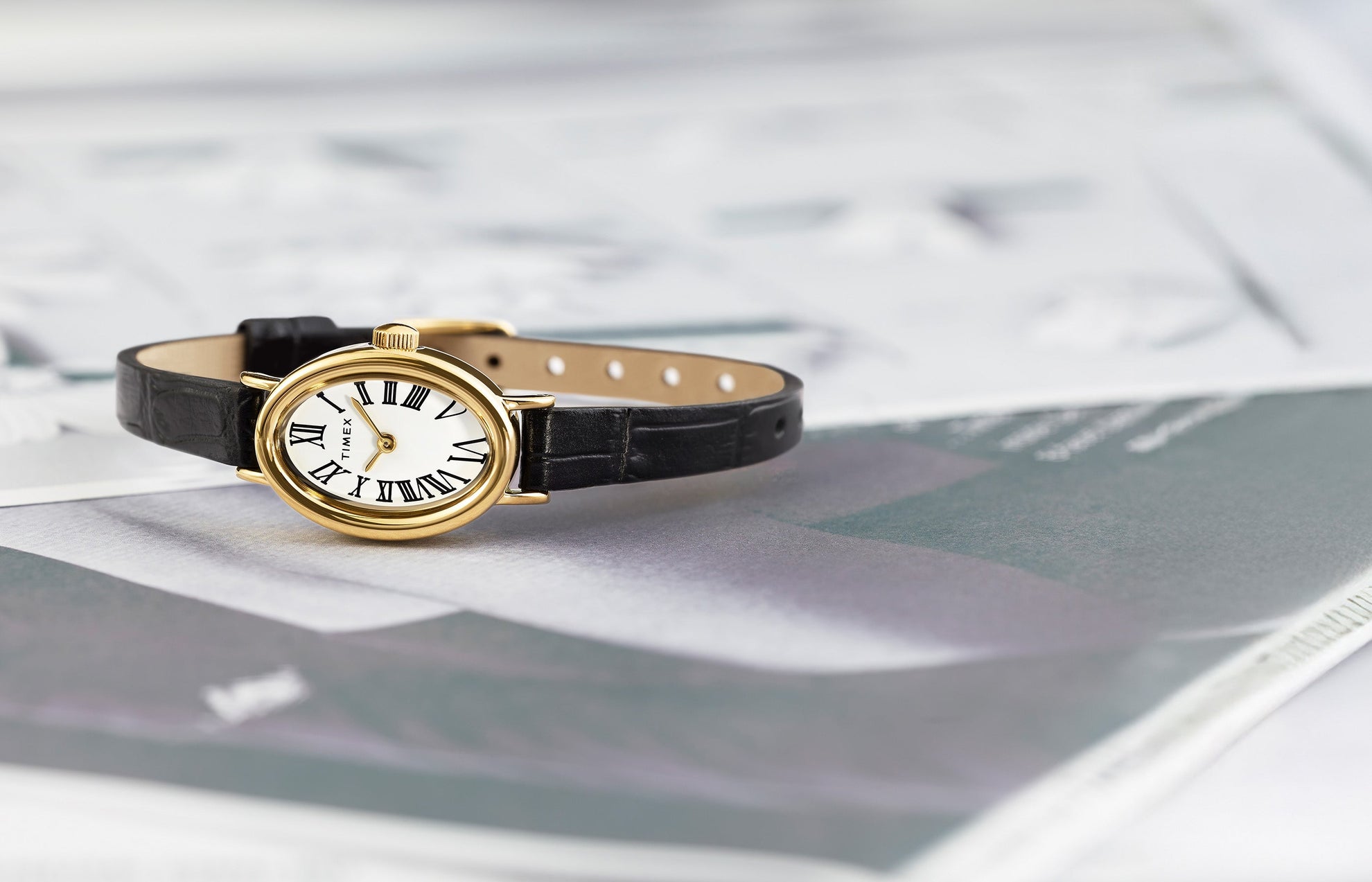 TIMEX® Cavatina watch with gold-tone oval case, white dial, and black leather strap resting on an open magazine.