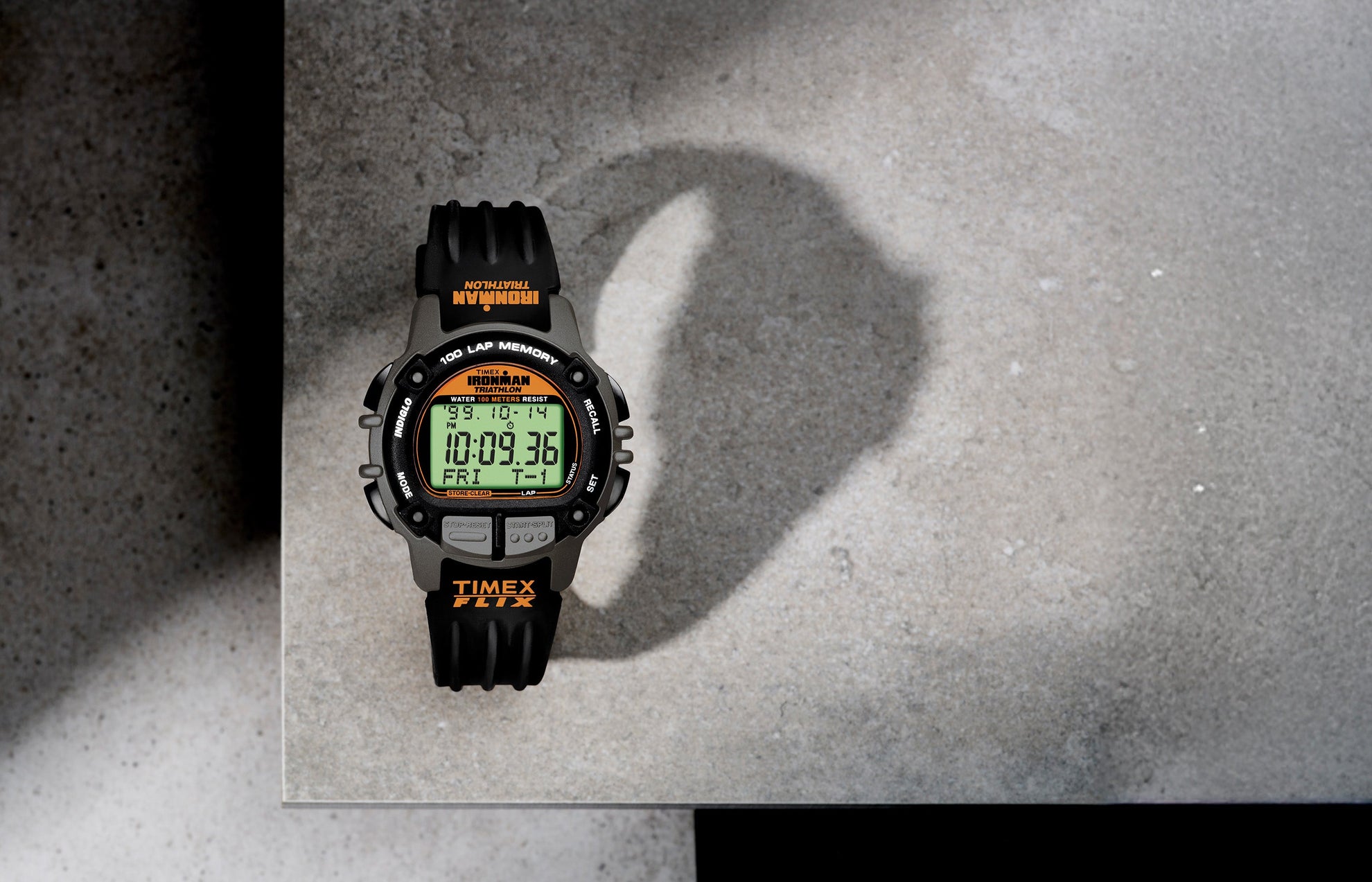 TIMEX® IRONMAN® Flix 100 digital sports watch with a black and gray case, orange accents, and green display, placed on a concrete surface casting a curved shadow.