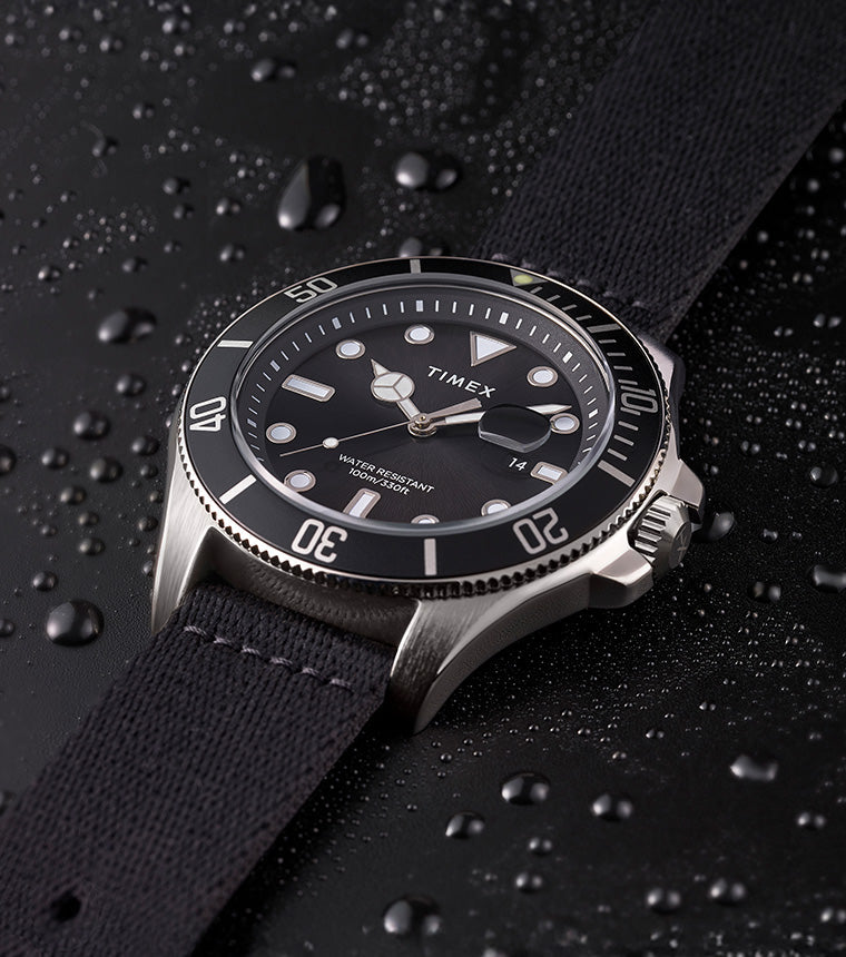 TIMEX® Harborside Coast watch with black dial and black fabric strap resting on a water-dappled black surface, showcasing its water-resistant design.