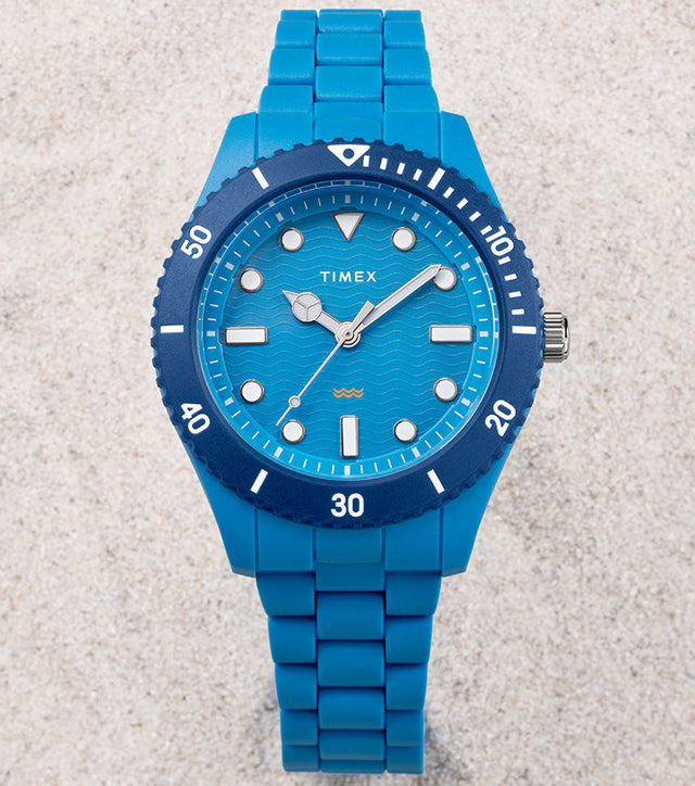 The TIMEX® Legacy Ocean #tide watch in bright blue rests on pale sand, featuring a wave-pattern dial and blue rotating bezel.