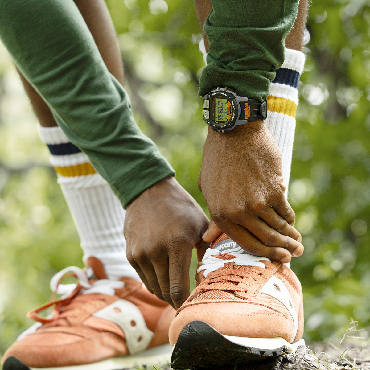 Runner wearing the TIMEX® IRONMAN® Flix 100 watch, tying orange sneakers in a green outdoor setting before a workout.
