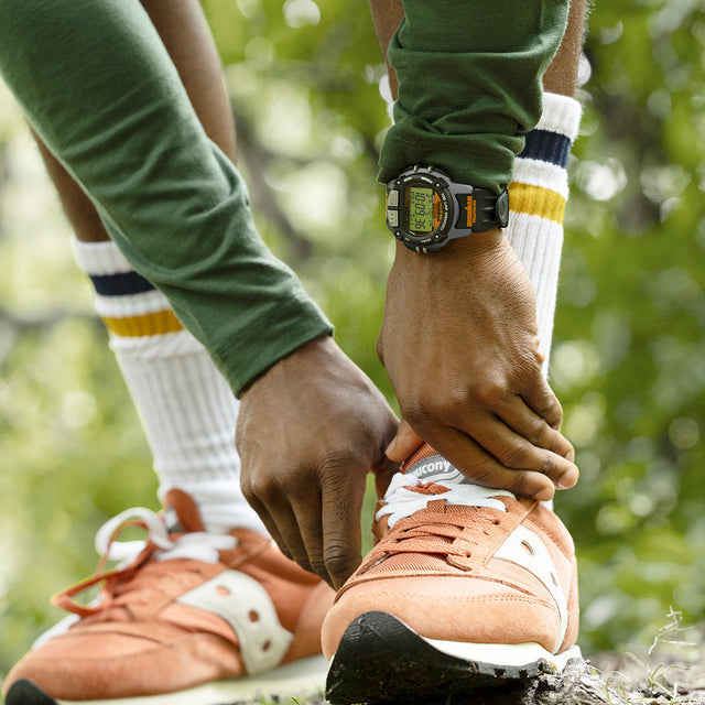Runner wearing the TIMEX® IRONMAN® Flix 100 watch, tying orange sneakers in a green outdoor setting before a workout.
