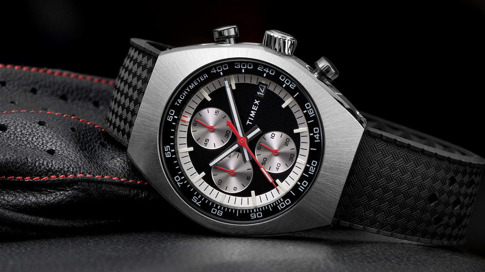 TIMEX® Legacy Tonneau Chronograph watch with black dial, silver-tone case, and textured black strap resting on perforated leather with red stitching.