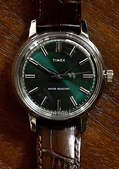 Todd Snyder x Timex Olive Marlin Automatic watch with a green sunray dial and brown leather strap, displayed flat on a textured wooden surface.