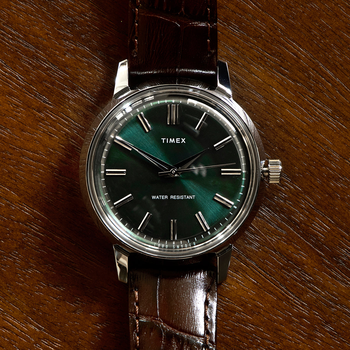 Front-facing view of the Todd Snyder x Timex Olive Marlin Automatic, showing its green dial, silver markers, and brown leather strap on a wood background.