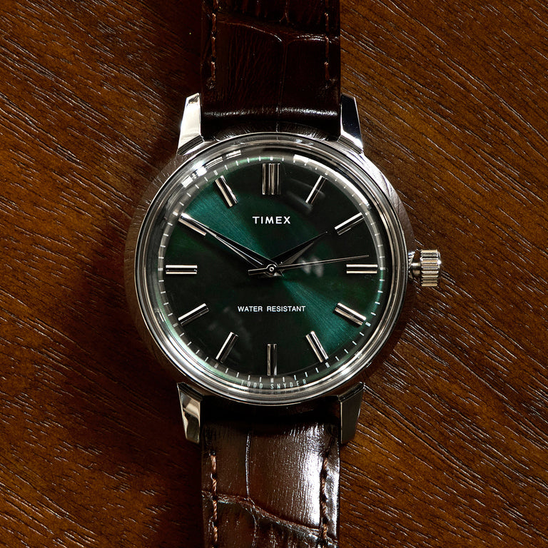 Front-facing view of the Todd Snyder x Timex Olive Marlin Automatic, showing its green dial, silver markers, and brown leather strap on a wood background.