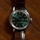 Front-facing view of the Todd Snyder x Timex Olive Marlin Automatic, showing its green dial, silver markers, and brown leather strap on a wood background.