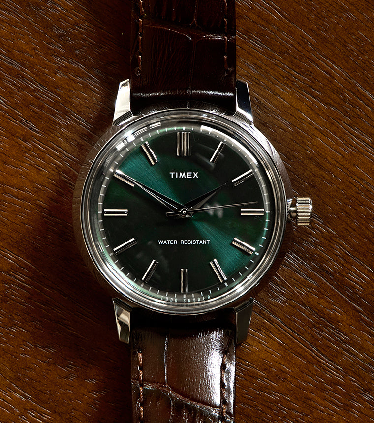 A Timex x Todd Snyder wristwatch with a deep green sunburst dial, silver hour markers, and a brown leather strap, shown resting on a textured wooden surface.
