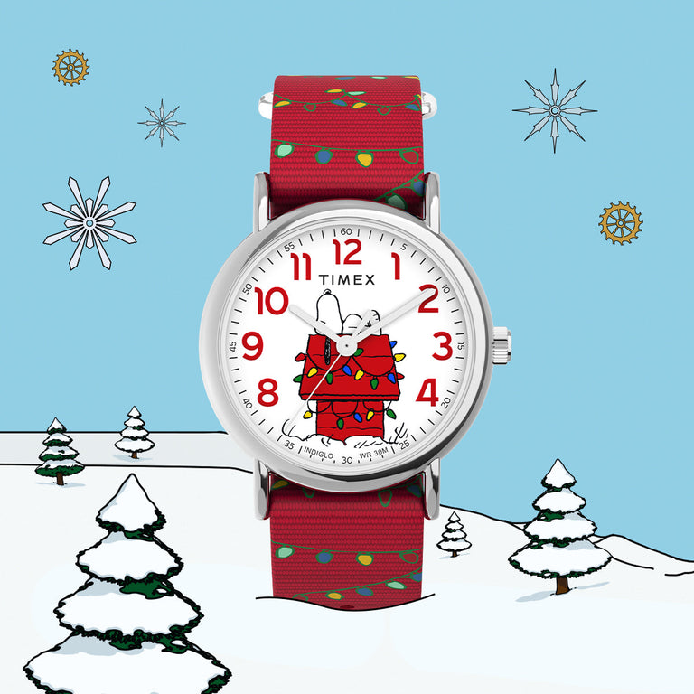 Timex x Peanuts holiday-themed watch featuring Snoopy wrapped in colorful Christmas lights on his red doghouse. The watch has a white dial with red numerals, a red fabric strap decorated with festive light patterns, and is set against a playful winter background with illustrated snow-covered trees and snowflakes.