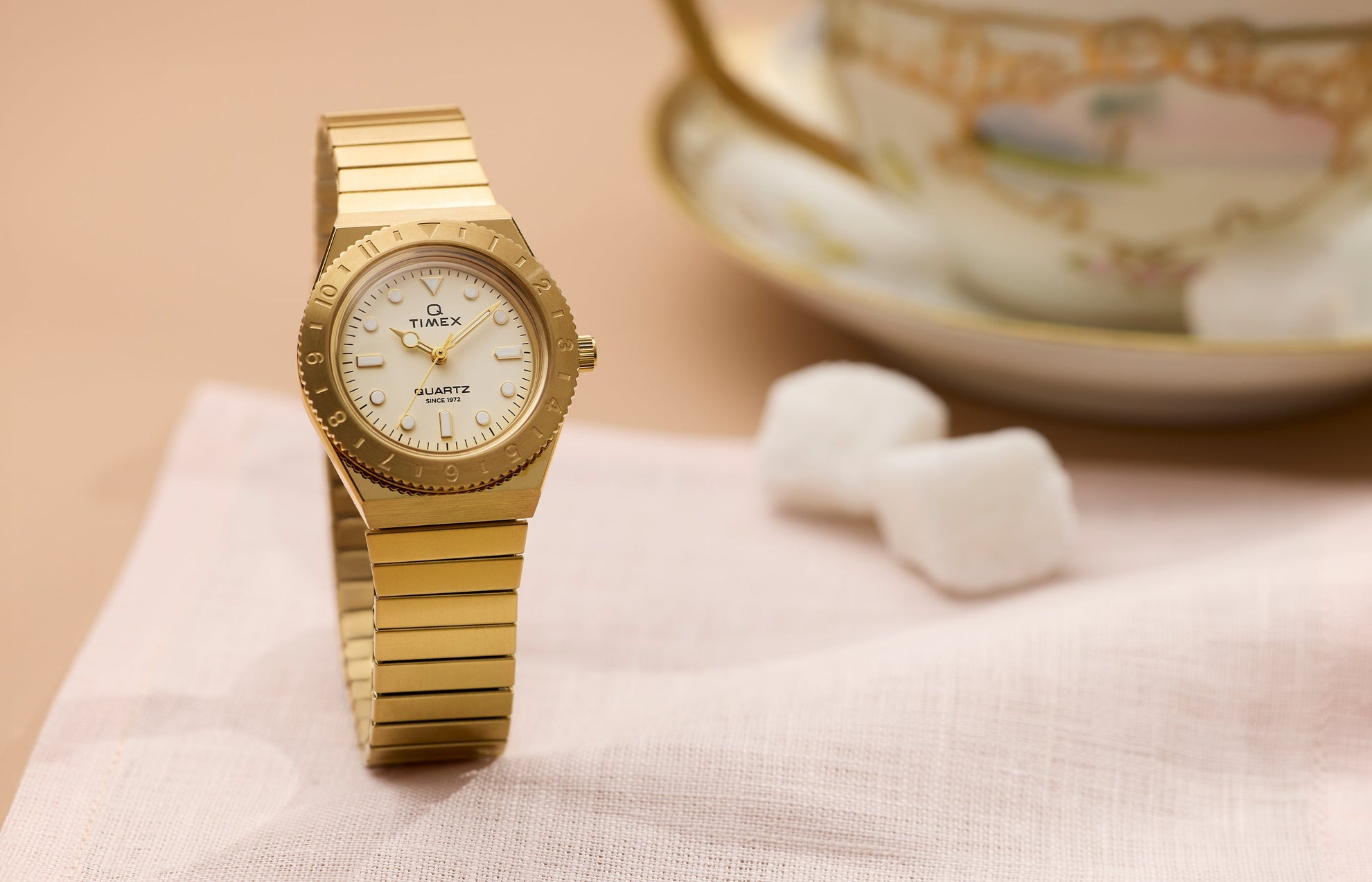 Q Timex® Mini gold-tone watch featuring a cream dial, rotating bezel, and gold expansion bracelet, displayed on a soft neutral tabletop.