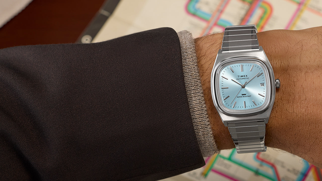 Timex® Automatic 1983 E Line watch with light blue dial and stainless steel bracelet worn on wrist, set against a vintage subway map background.