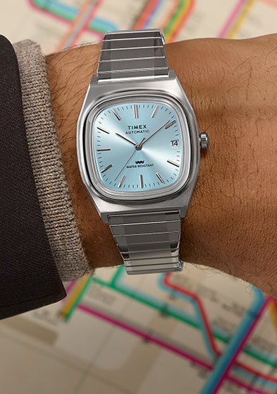 Timex® Automatic 1983 E Line watch with light blue dial and stainless steel bracelet worn on wrist, set against a vintage subway map background.