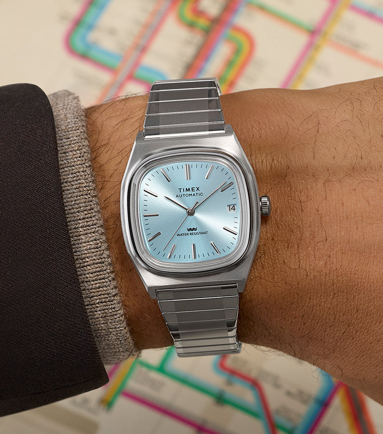 Timex® Automatic 1983 E Line watch with a light blue dial and stainless steel bracelet worn on wrist, featuring a day-date window and brushed silver-tone case, shown against a colorful transit map background.