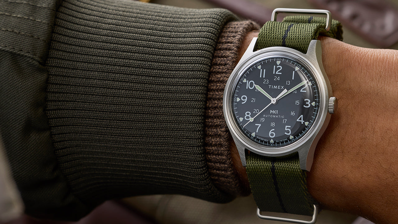 Timex MK1 Automatic worn on wrist with green fabric strap, stainless steel case, and black dial with Arabic numerals.