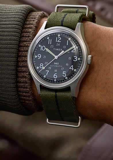 Timex MK1 Automatic worn on wrist with green fabric strap, stainless steel case, and black dial with Arabic numerals.