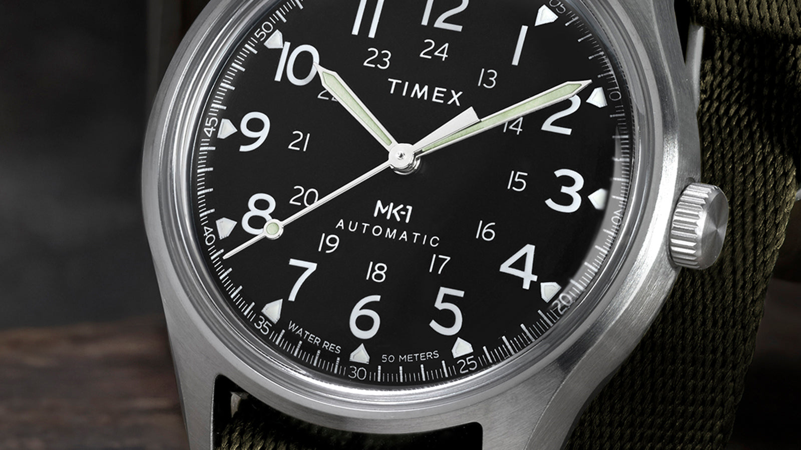 Macro view of Timex MK1 Automatic dial showing luminous hands, minute markers, and 24-hour military time scale.