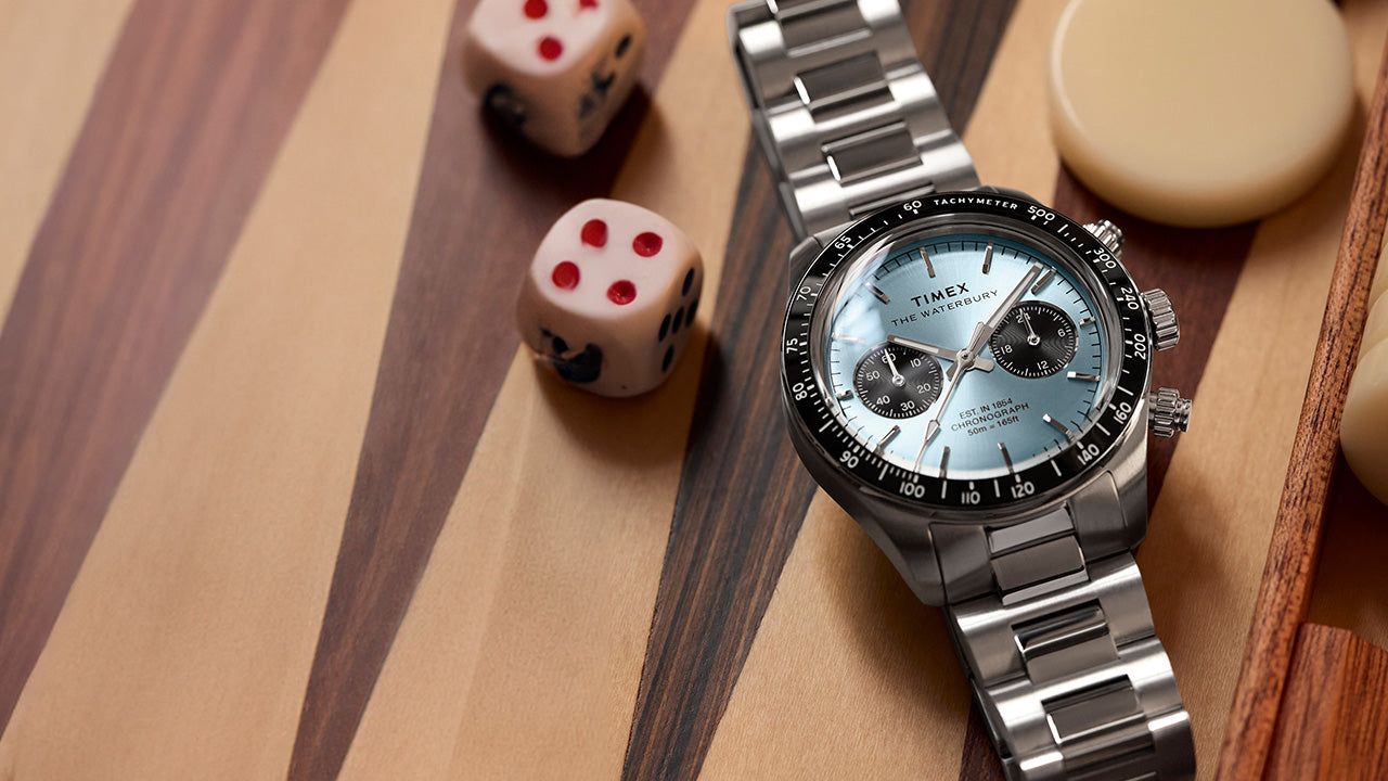 Timex Waterbury Heritage Chronograph with stainless steel bracelet and light blue dial, displayed on a wooden game board with dice.