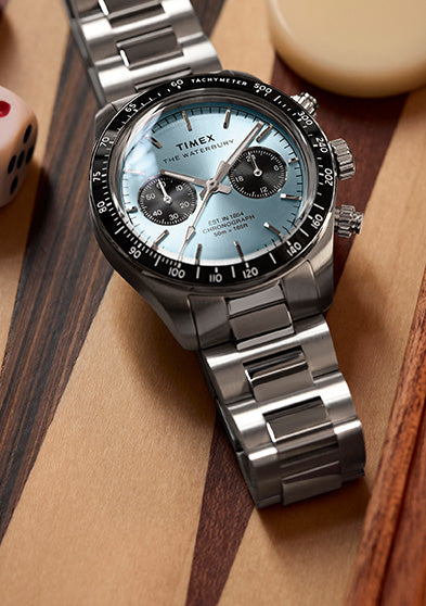 Timex Waterbury Heritage Chronograph with stainless steel bracelet and light blue dial, displayed on a wooden game board with dice.