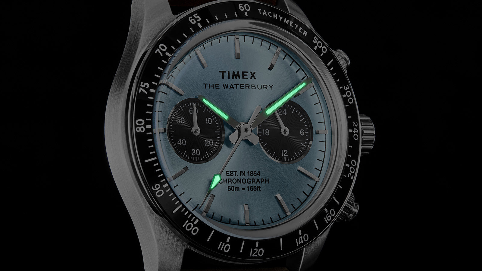 Timex Waterbury Heritage Chronograph dial in low light, highlighting luminous hands and hour markers on a light blue dial.