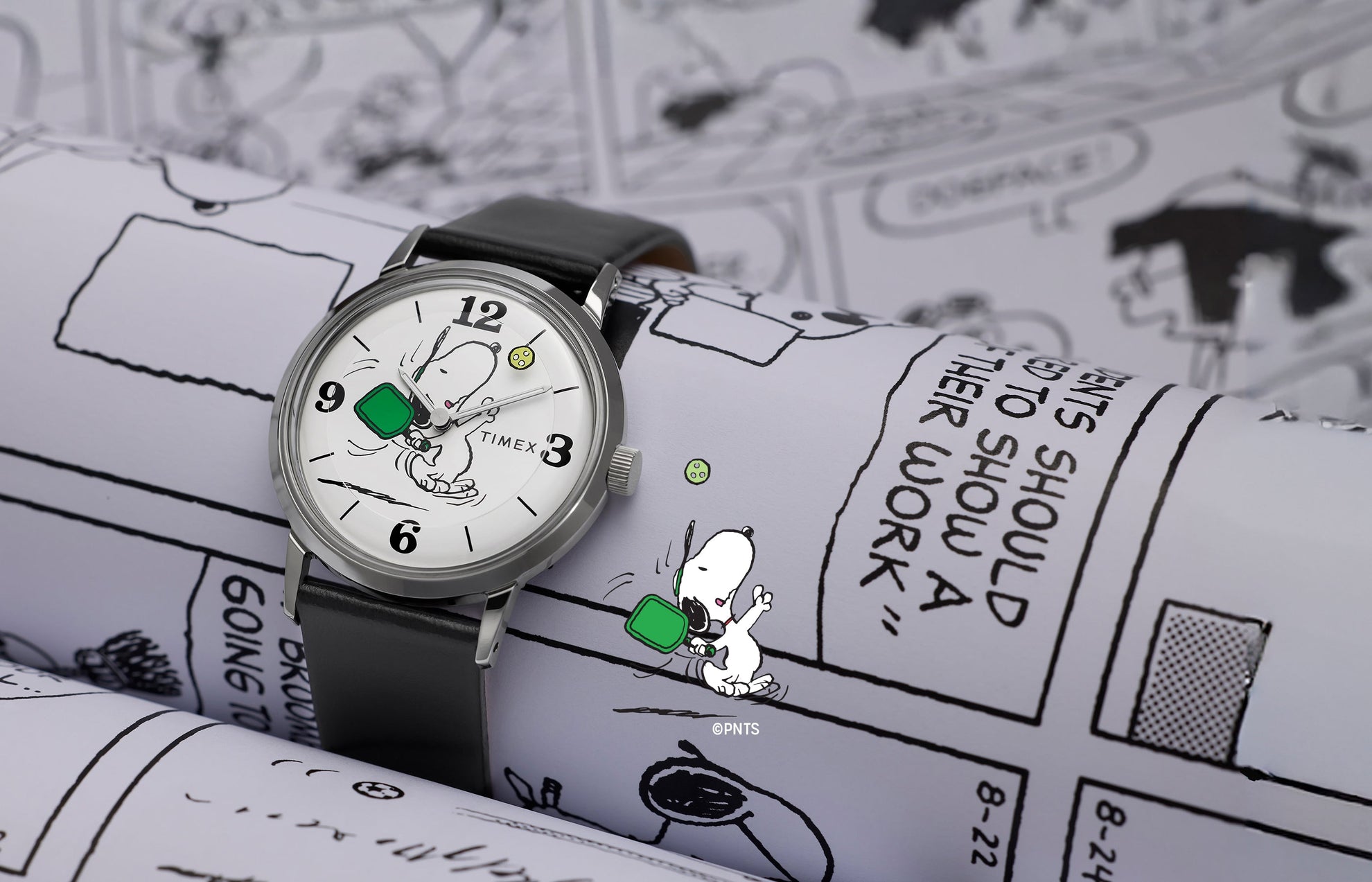 Peanuts® x Timex Marlin® Pickleball watch with black leather strap and white dial featuring Snoopy playing pickleball, displayed on Peanuts comic strip background.