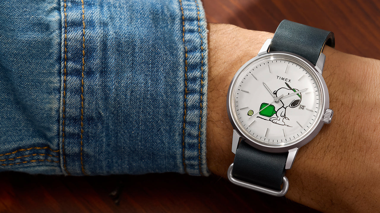 Peanuts® x Timex Marlin® Automatic Pickleball watch worn on wrist, featuring Snoopy playing pickleball on a white dial, stainless-steel case, date window, and dark green leather strap.