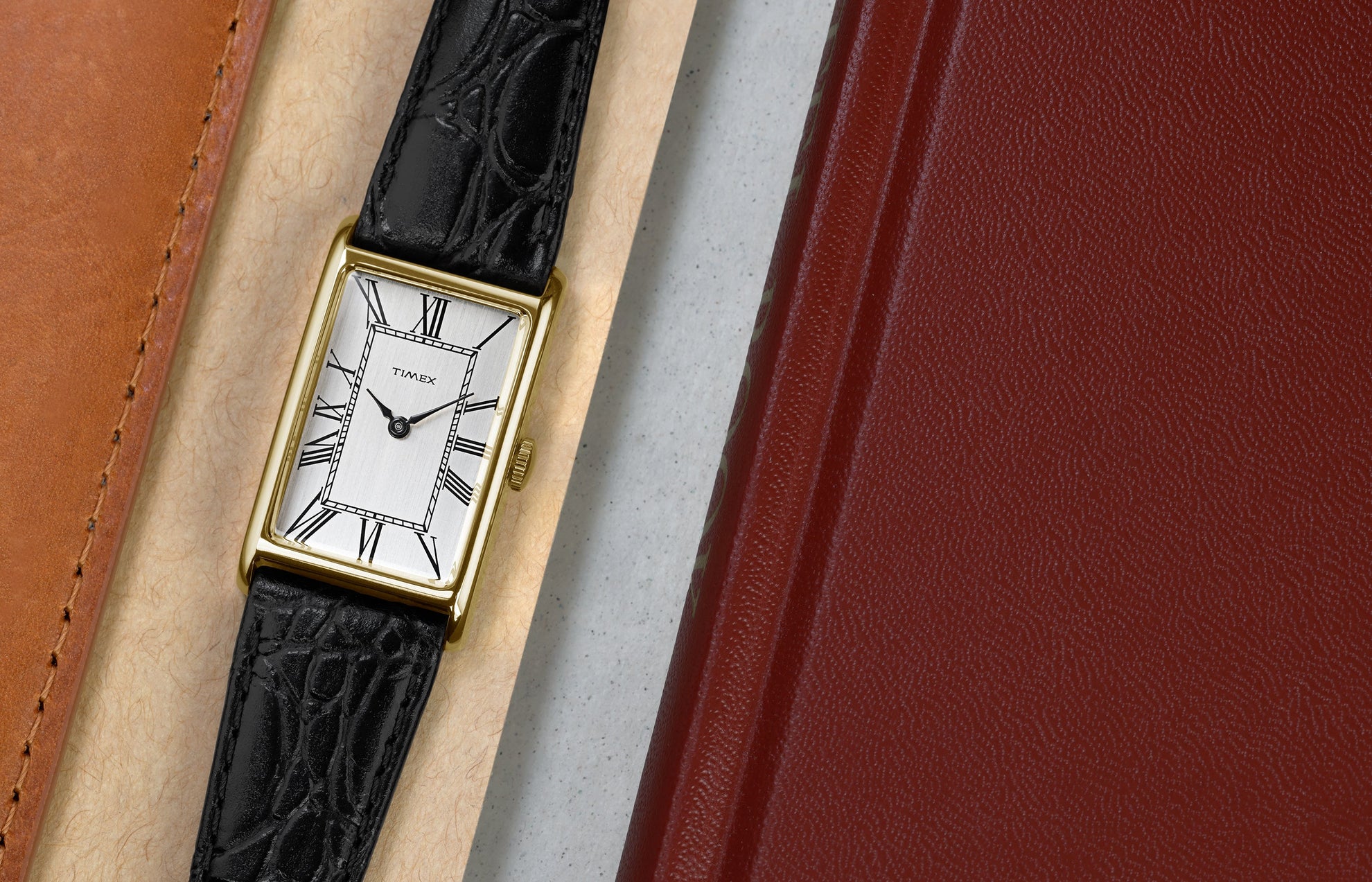Timex 1976 Lexington Reissue watch laid flat beside leather accessories, highlighting the slim gold-tone case and classic Roman numeral dial.