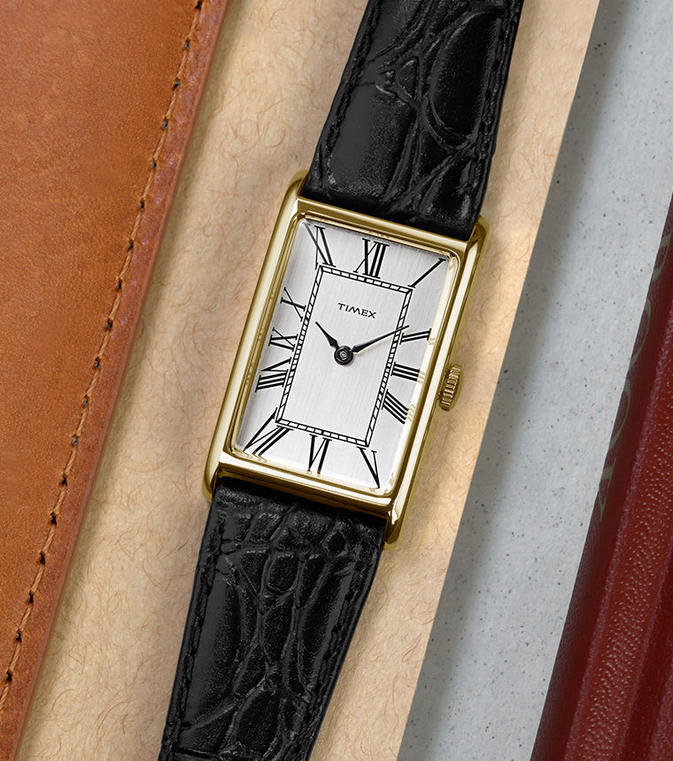 Timex 1976 Lexington Reissue watch laid flat beside leather accessories, highlighting the slim gold-tone case and classic Roman numeral dial.