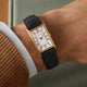 Gold-tone rectangular Timex 1976 Lexington Reissue watch with a silver dial and black Roman numerals, worn on a wrist with a black embossed leather strap.