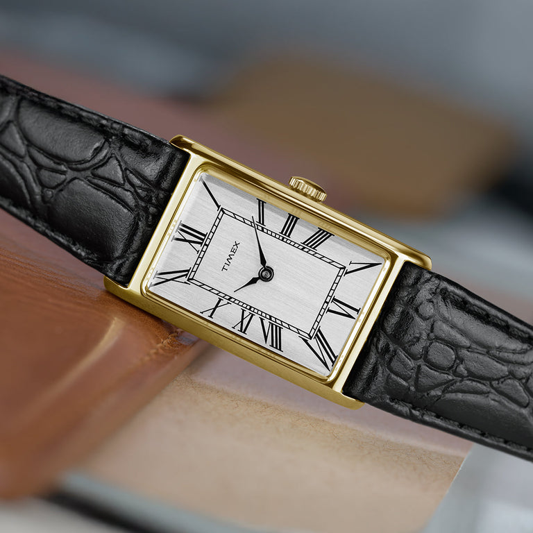 Close-up of the Timex 1976 Lexington Reissue featuring a gold-tone rectangular case, brushed silver dial, black Roman numerals, and black leather strap resting on a leather surface.