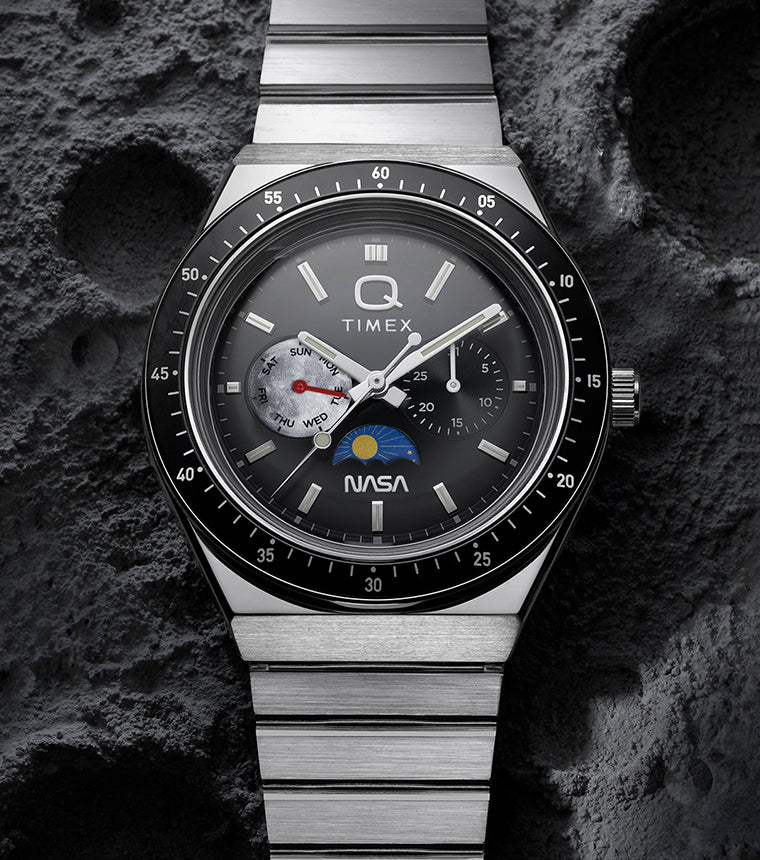 Q Timex® x NASA stainless steel watch with black bezel and multifunction gray dial displayed against a textured lunar surface background.