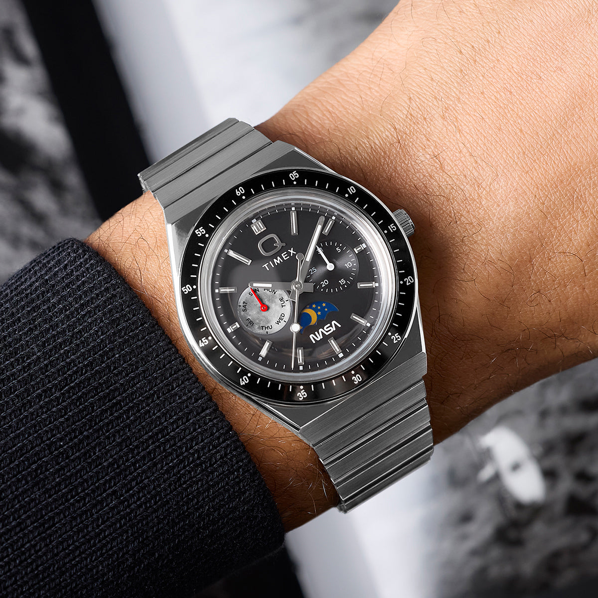 Q Timex® x NASA watch worn on wrist, showcasing stainless steel bracelet, black timing bezel, gray multifunction dial, and NASA logo detail.