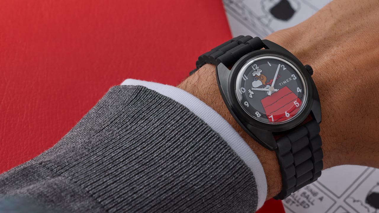 Peanuts® x Timex 1979 Reissue Inspired watch worn on wrist, featuring a black case and bracelet with Snoopy as the Flying Ace on a gray dial above his red doghouse.