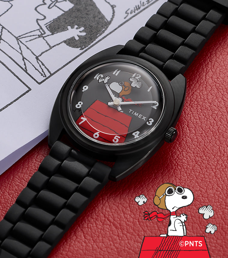 Peanuts® x Timex 1979 Reissue Inspired watch with black bracelet and gray dial showcasing Snoopy Flying Ace artwork, placed on a red notebook beside Peanuts comic strip pages.