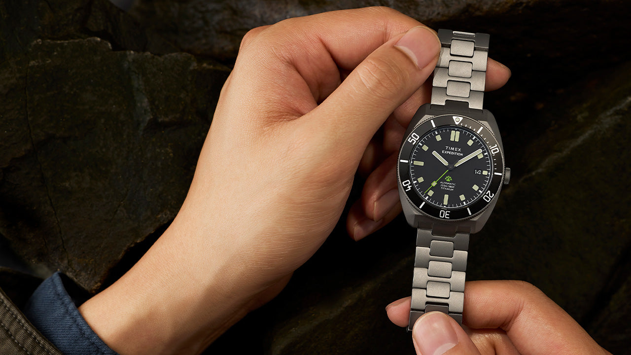 Expedition® Pioneer Titanium Automatic watch held in hand, featuring a brushed titanium case and bracelet, black dial, and green seconds hand.
