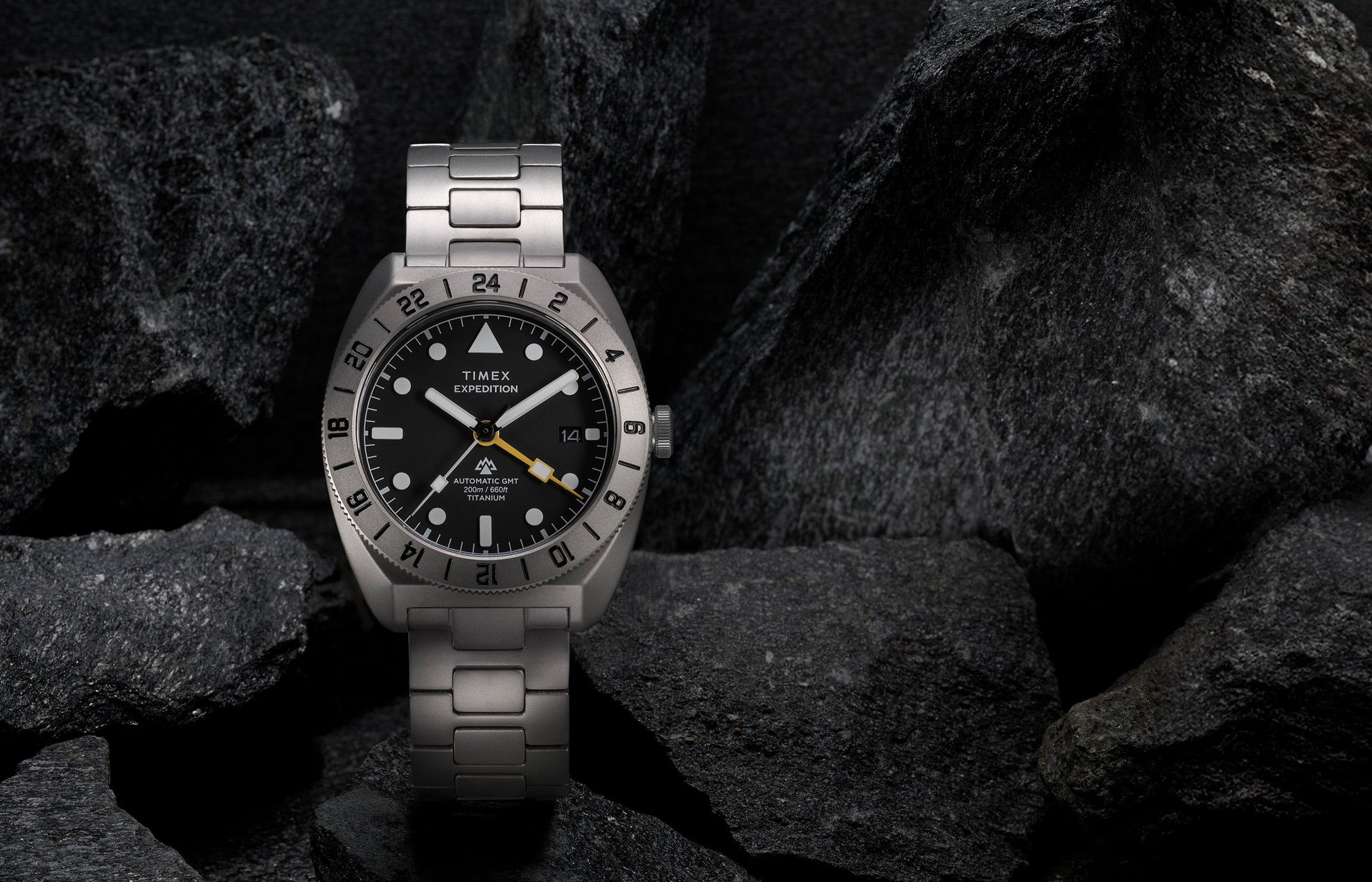 Expedition® Pioneer Titanium Automatic GMT standing upright on rocks, showcasing the titanium bracelet, black dial, and engraved 24-hour bezel.