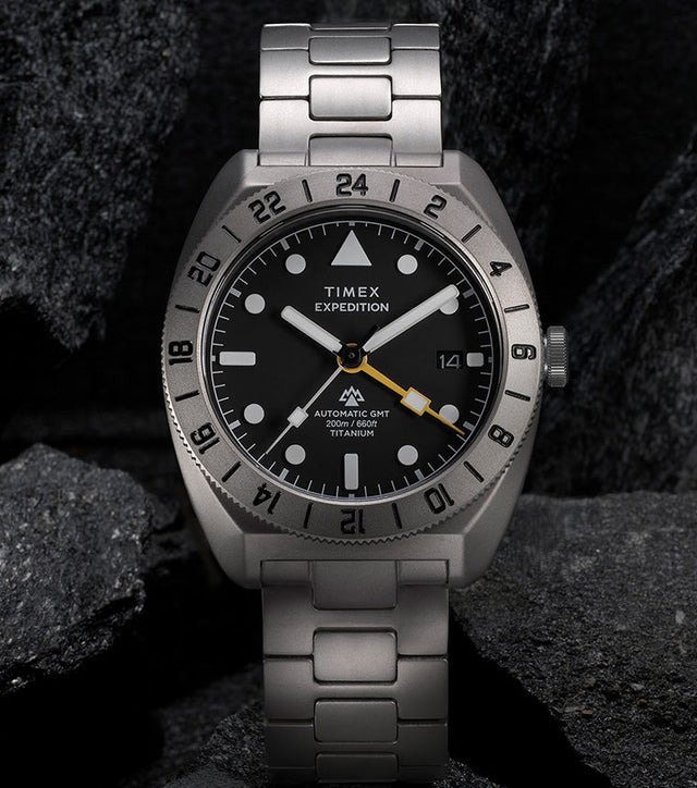Expedition® Pioneer Titanium Automatic GMT standing upright on rocks, showcasing the titanium bracelet, black dial, and engraved 24-hour bezel.