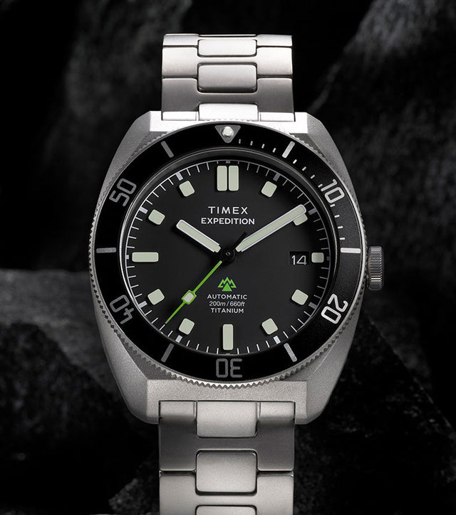 Expedition® Pioneer Titanium Automatic standing on rugged rocks, highlighting the titanium bracelet, black dial, rotating bezel, and date window.