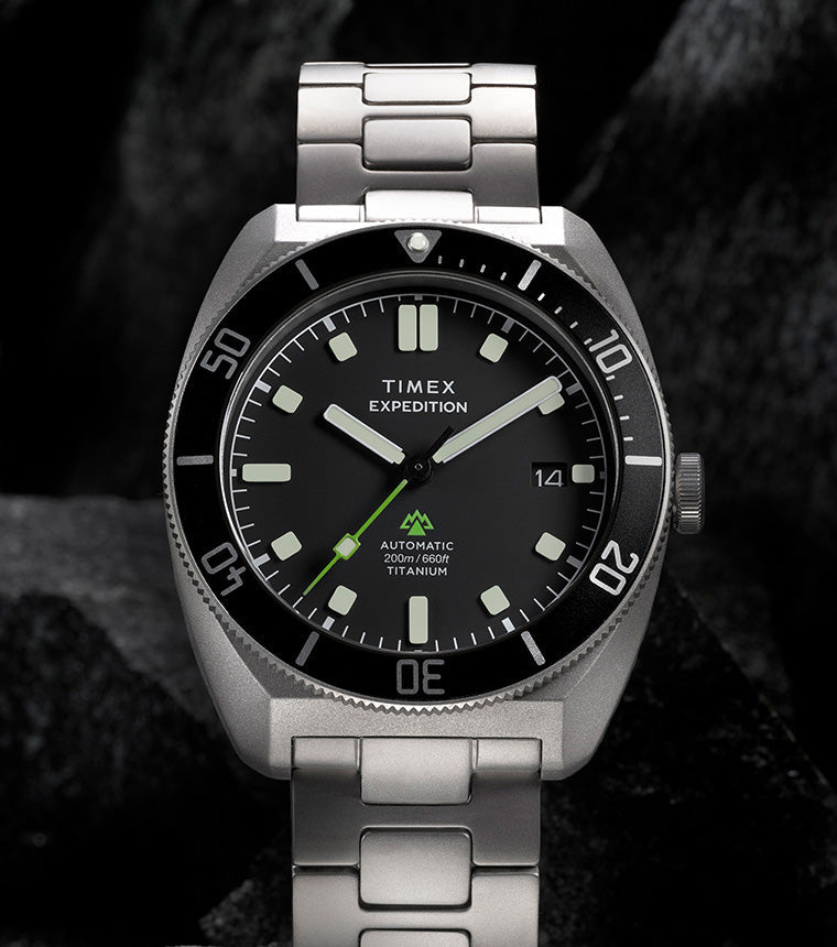 Expedition® Pioneer Titanium Automatic standing on rugged rocks, highlighting the titanium bracelet, black dial, rotating bezel, and date window.